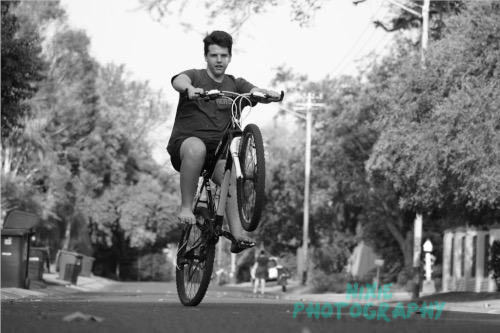 A black and white photo of a boy doing a wheelie on his bicycle in the street
